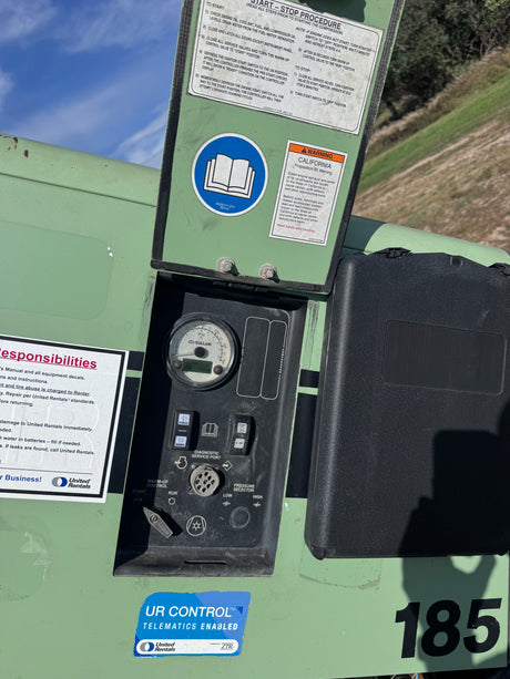 Close-up of a green industrial control panel with an open lid showing manuals and stickers, a black control box, and the number 185 on a machine outdoors.