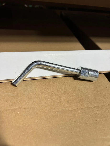 A silver metal hex wrench with a bent shaft and hexagonal head, resting on a brown cardboard surface next to a white board.