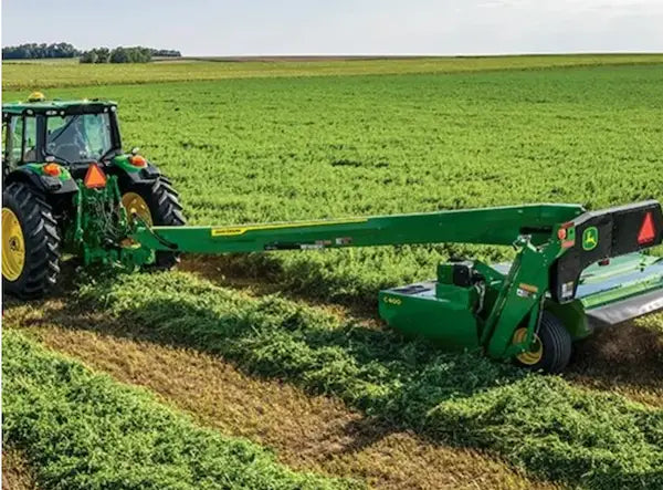 A green John Deere tractor pulling a large green hay cutter or baler attachment across a wide green field.